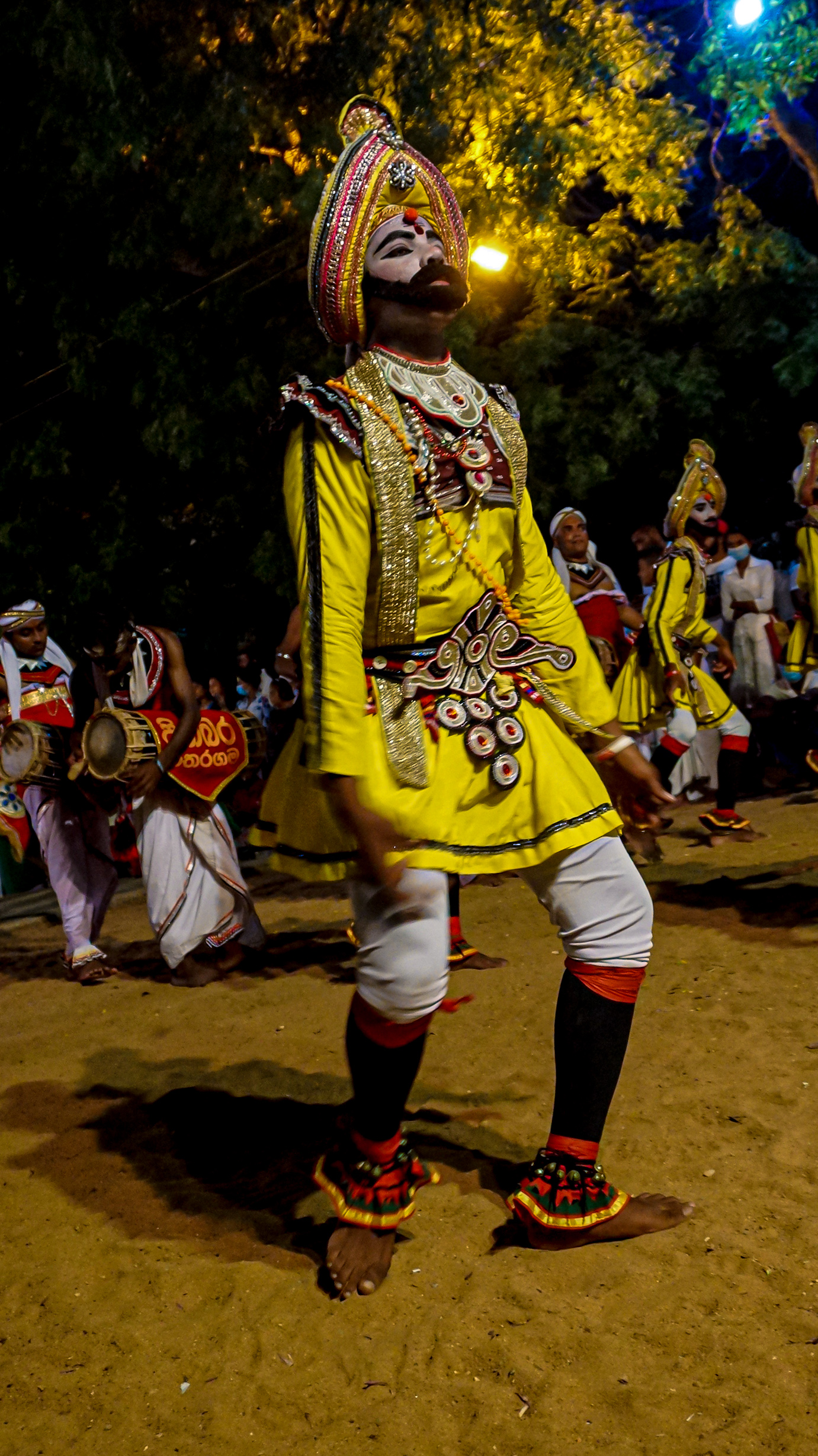 A Wadiga Patuna dancer in a brilliant yellow costume and headdress, with drummers behind him at night.